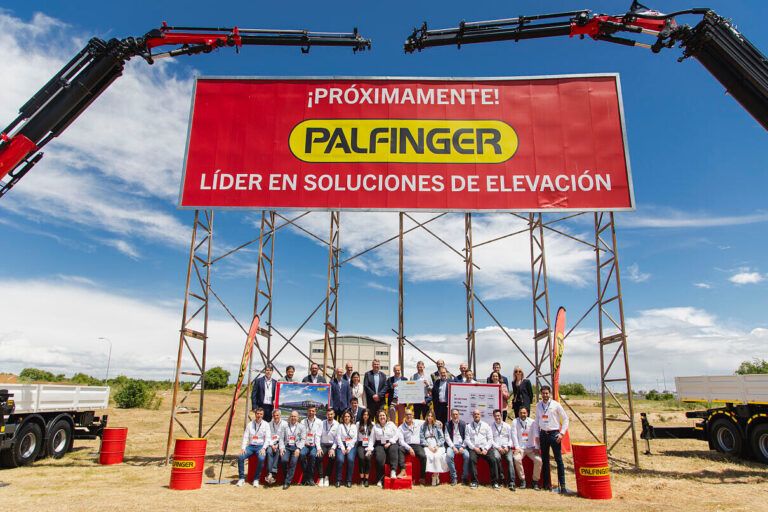 Photograph of Palfinger employees gathered beneath a large red Palfinger banner, flanked by two truck-mounted Palfinger cranes raised to form an arch over the group.