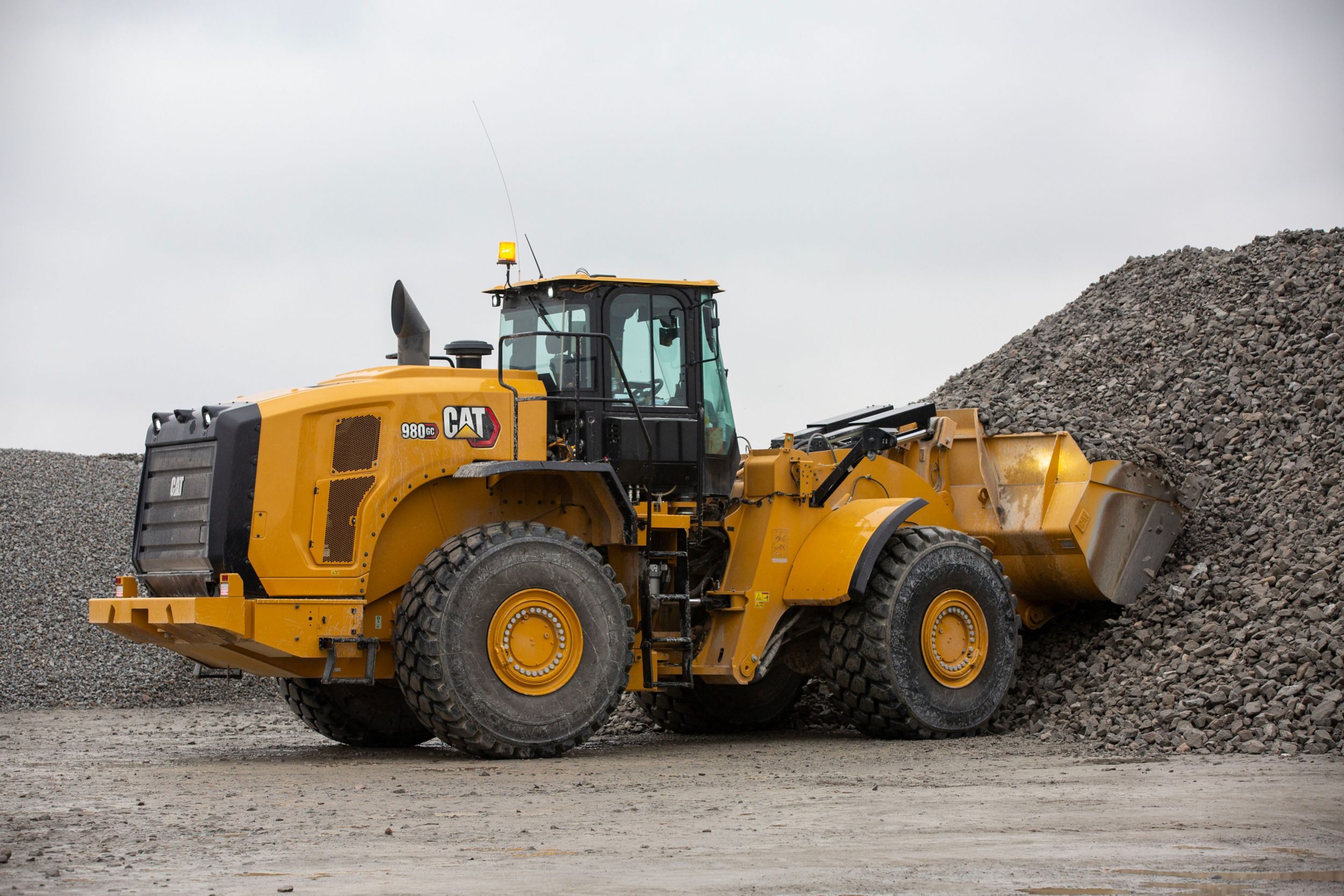 The Cat 980 GC wheel loader is shown at a construction or mining site. The loader is positioned next to a large pile of gravel or rocks, with its bucket partially filled and raised