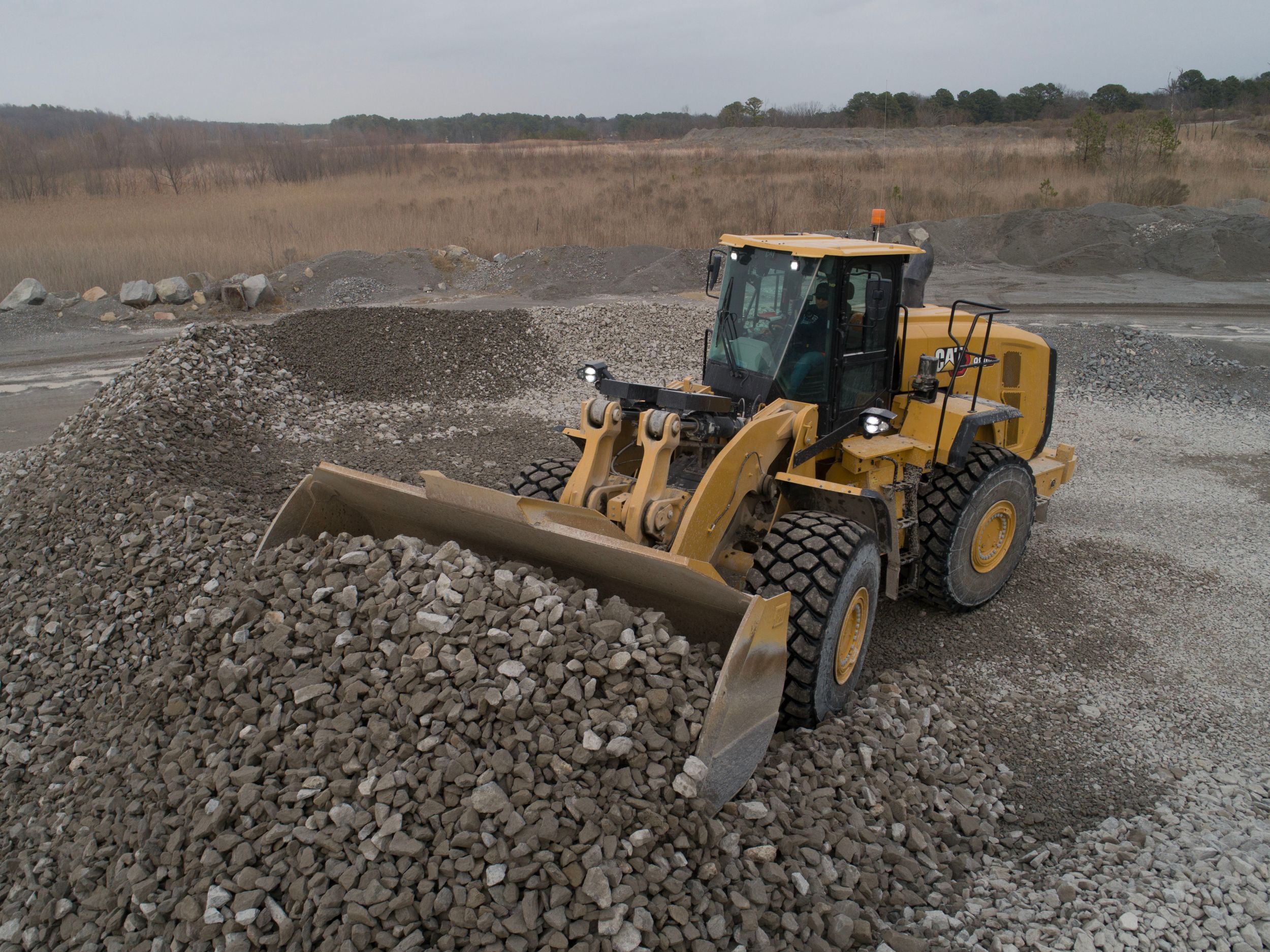 The Cat 980 GC wheel loader is shown in an outdoor construction or quarry site. The loader's bucket is filled with a pile of gravel or small rocks