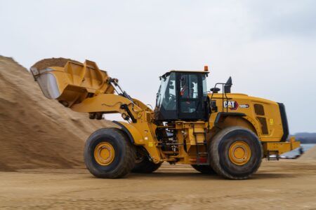 The Cat 980 GC wheel loader is in motion on a construction site, carrying a load of dirt in its front bucket. The machine is positioned against a backdrop of a large mound of dirt and an overcast sky