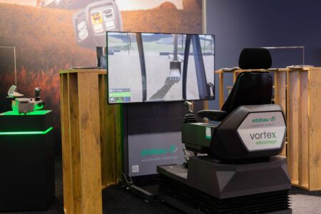 Elobau to showcase control and sensor technology at ConExpo An elobau exhibition stand featuring the vortex advantage operator simulator, consisting of a black seat with armrest-mounted joysticks facing a large screen displaying a virtual excavator environment. Additional control elements are displayed on a lit plinth to the left.