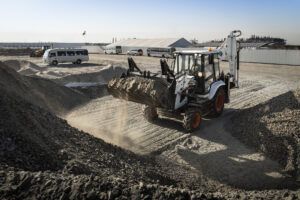 A white and black Bobcat B730 M-Series backhoe loader with orange wheels moving through gravel stockpiles on a dusty construction site, with minibuses and industrial buildings visible in the background.