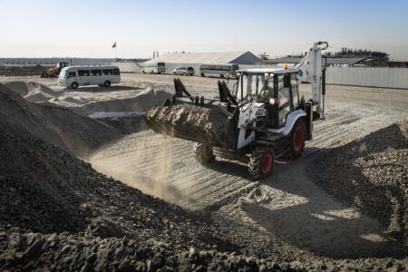 Bobcat introduces AutoShift transmission for B730 M-Series backhoe loader A white and black Bobcat B730 M-Series backhoe loader with orange wheels moving through gravel stockpiles on a dusty construction site, with minibuses and industrial buildings visible in the background.