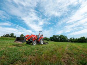 Case IH Farmall compact tractor with front loader bucket operating in an open field, with rear-mounted mower attachment