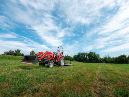 Case IH Farmall compact tractor with front loader bucket operating in an open field, with rear-mounted mower attachment