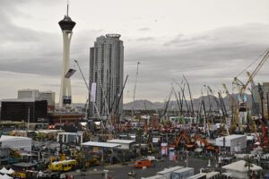 An elevated view of the ConExpo-Con/Agg outdoor exhibition area in Las Vegas, with rows of construction equipment, cranes and branded manufacturer booths stretching into the distance, and the Strat hotel tower visible on the left against an overcast sky.