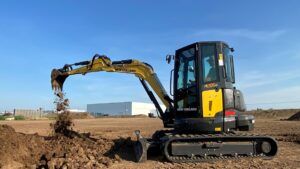 A New Holland E50D D-Series mini excavator working on a flat construction site, its yellow arm raised and bucket releasing excavated soil. An operator is visible in the enclosed cab.