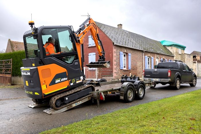 A Develon DX23E-7 electric mini-excavator being loaded onto a twin-axle trailer towed by a pickup truck on a village road.