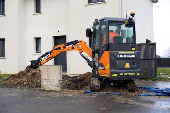 A Develon DX23E-7 electric mini-excavator digging beside a residential building, with an operator visible in the enclosed cab