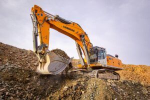 A Liebherr 992 crawler excavator in orange livery operates on a rocky quarry face, its bucket engaged in material excavation against an overcast sky.