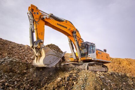 Liebherr outlines digital roadmap with ConExpo showcase A Liebherr 992 crawler excavator in orange livery operates on a rocky quarry face, its bucket engaged in material excavation against an overcast sky.