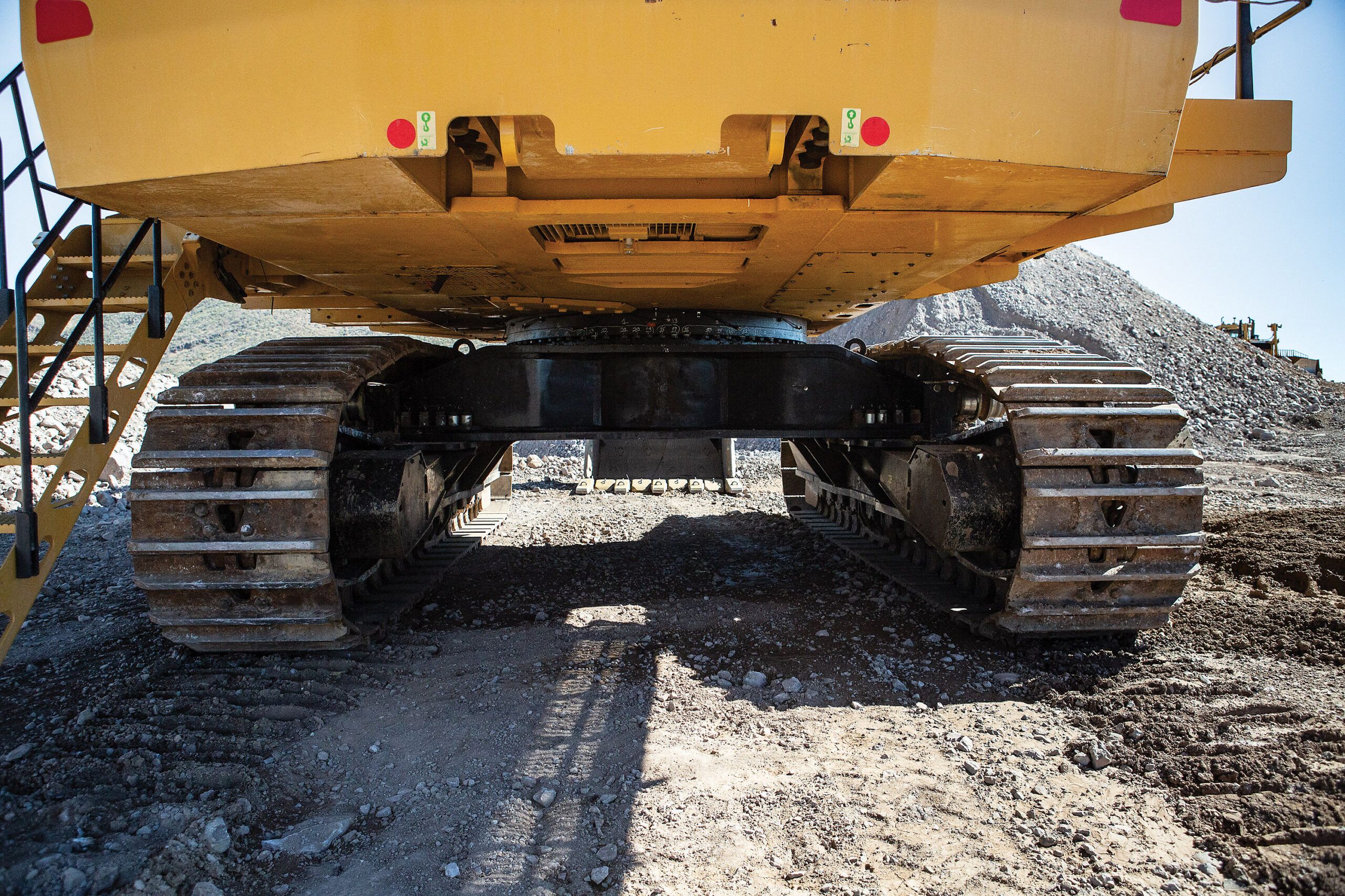 Low-angle view of Cat 6015 hydraulic mining shovel's undercarriage showing twin heavy-duty track assemblies, protective belly guards and access stairway in a quarry setting.