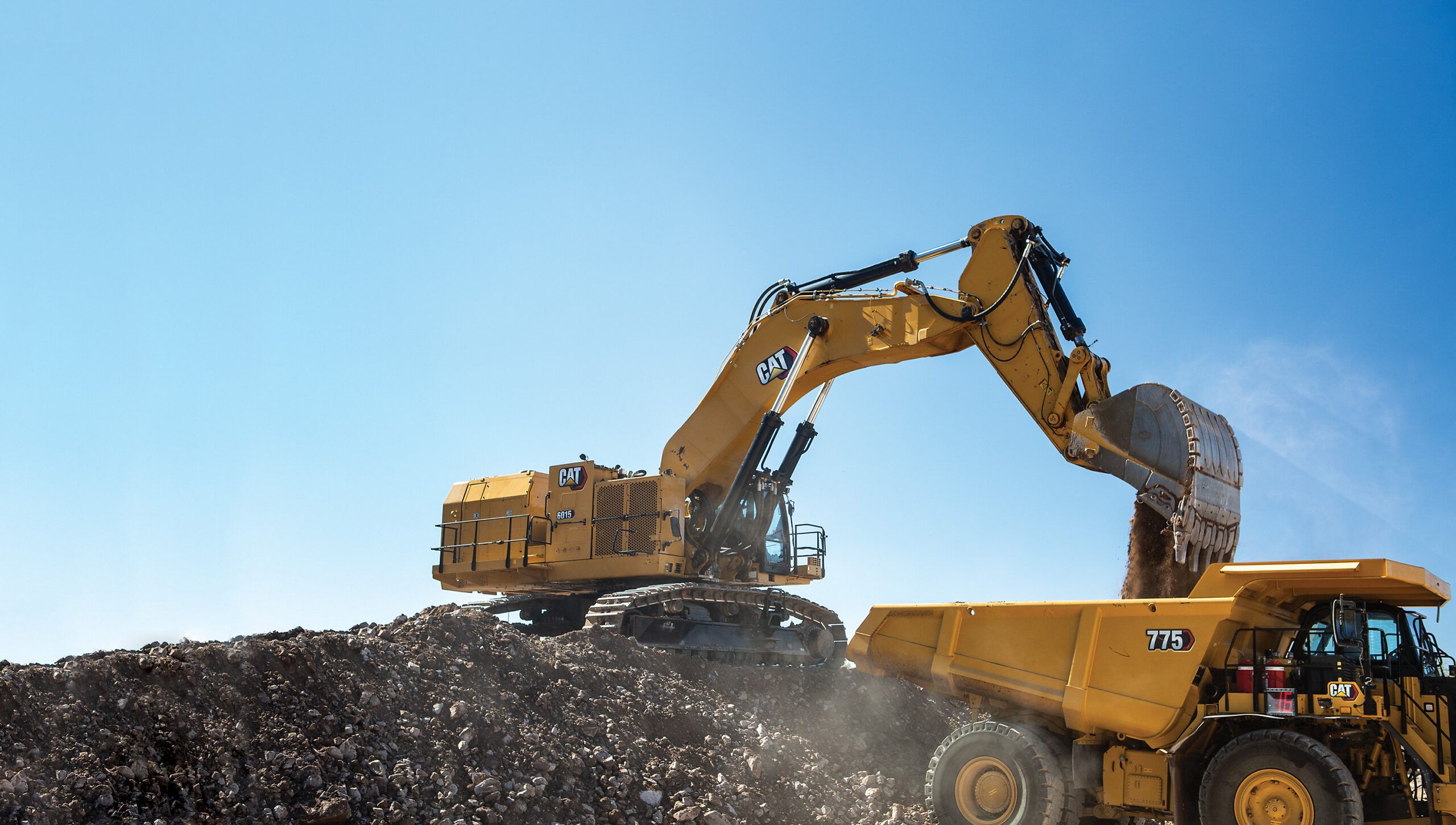 Cat 6015 hydraulic mining shovel loading material into a Cat 775 haul truck from an elevated position at a mining site.