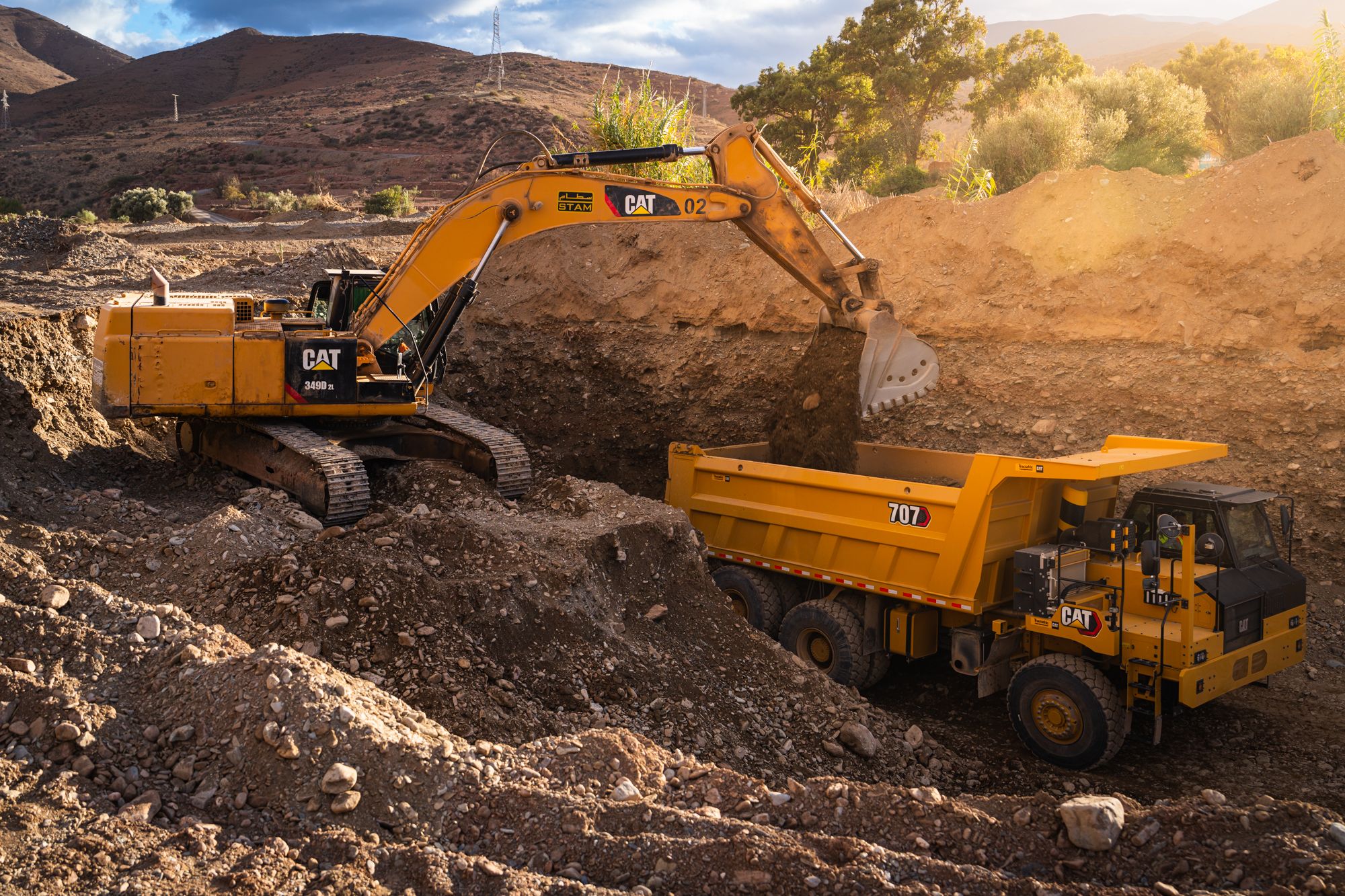 A Cat 349D XE excavator loads material into a Cat 707 wide body truck at an earthmoving site, with arid hillside terrain visible in the background.