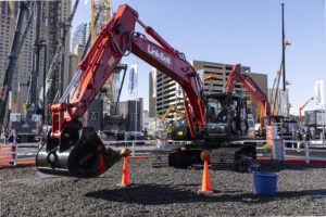 A red Link-Belt E400 crawler excavator on a gravel demonstration area at an outdoor trade show, with an operator in the cab. Orange traffic cones and basketballs are positioned around the machine as part of a skills demonstration. Cranes, equipment and city buildings are visible in the background.