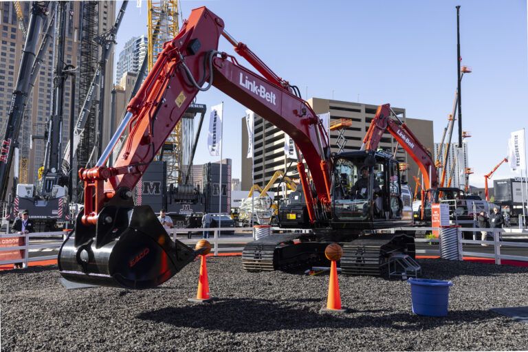 A red Link-Belt E400 crawler excavator on a gravel demonstration area at an outdoor trade show, with an operator in the cab. Orange traffic cones and basketballs are positioned around the machine as part of a skills demonstration. Cranes, equipment and city buildings are visible in the background.