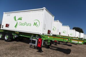 A line of white GeoPura hydrogen transport containers bearing the GeoPura H2 logo in green, mounted on green-framed flatbed trailers under a blue sky.