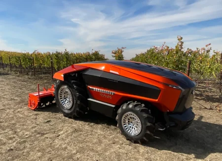 The Kubota Agri Concept autonomous agricultural robot, finished in orange and black, positioned in a vineyard with a red tilling attachment fitted to its rear. The cabless, low-profile machine sits on four large agricultural tyres against a backdrop of vine rows and a partly cloudy sky.