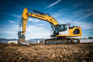A yellow and white Liebherr 924 crawler excavator digging into rocky ground on a construction site, with mountains and a clear blue sky in the background.