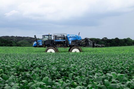 New Holland launches updated defensor self-propelled sprayer range A blue New Holland defensor self-propelled sprayer travelling through a green crop field with booms folded, under an overcast sky