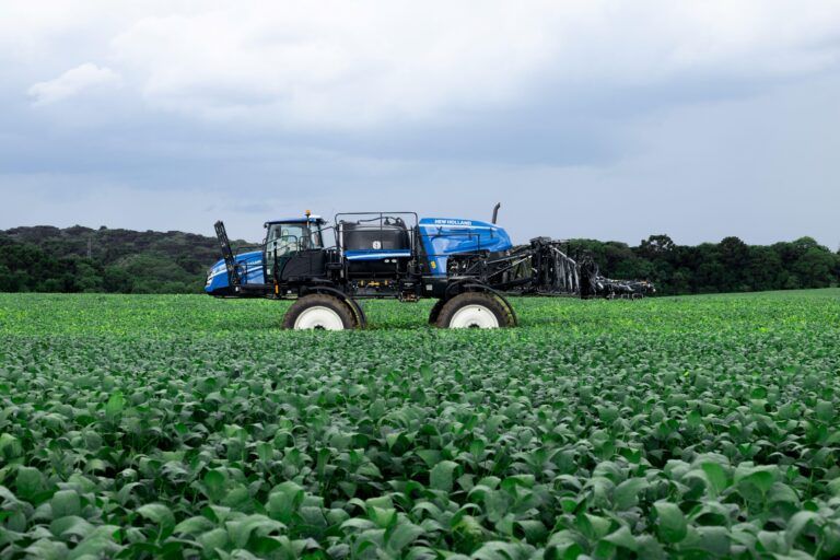 A blue New Holland defensor self-propelled sprayer travelling through a green crop field with booms folded, under an overcast sky