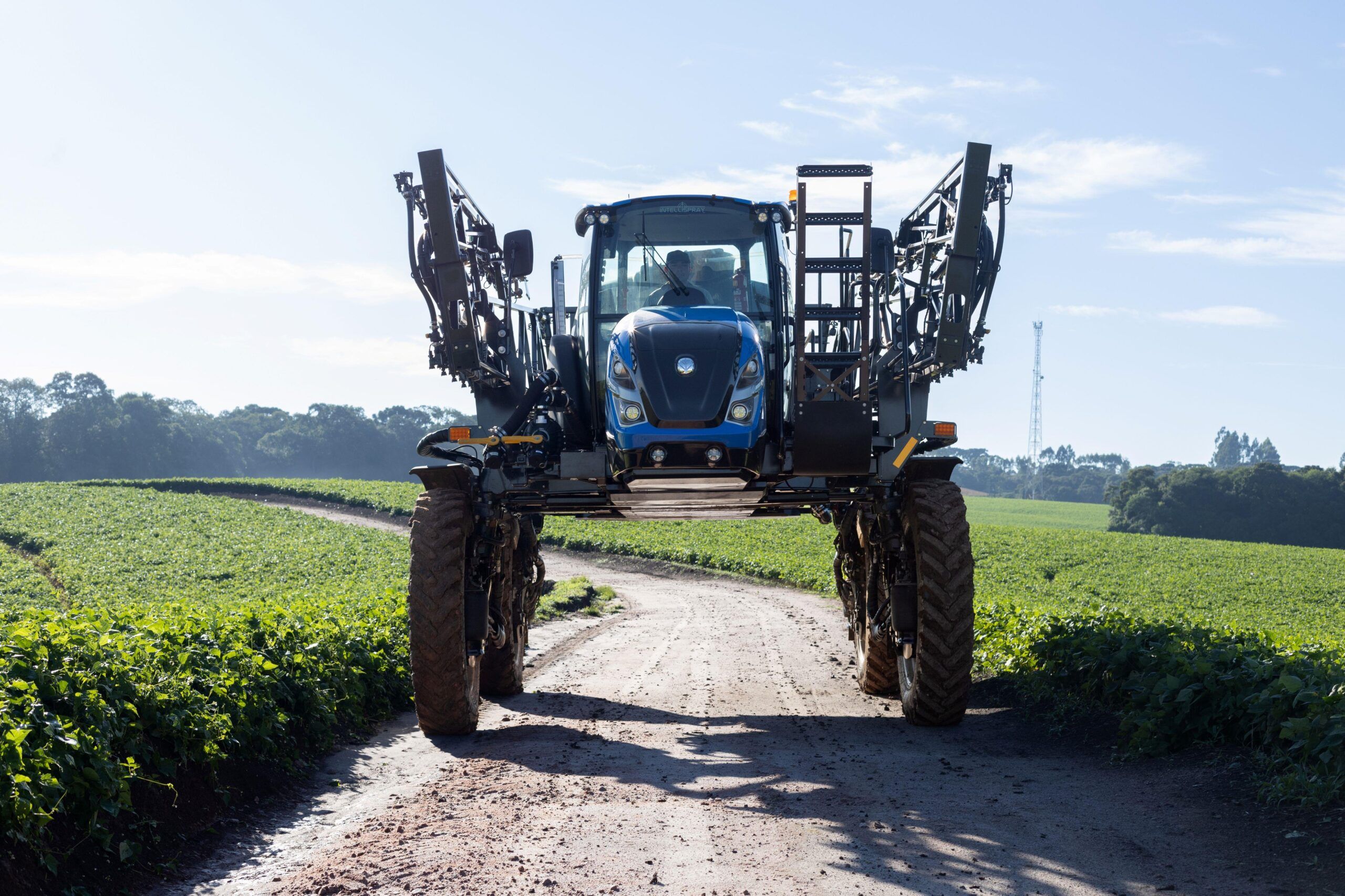 A blue New Holland defensor self-propelled sprayer viewed from the front with booms fully extended, positioned on a dirt track between green crop fields