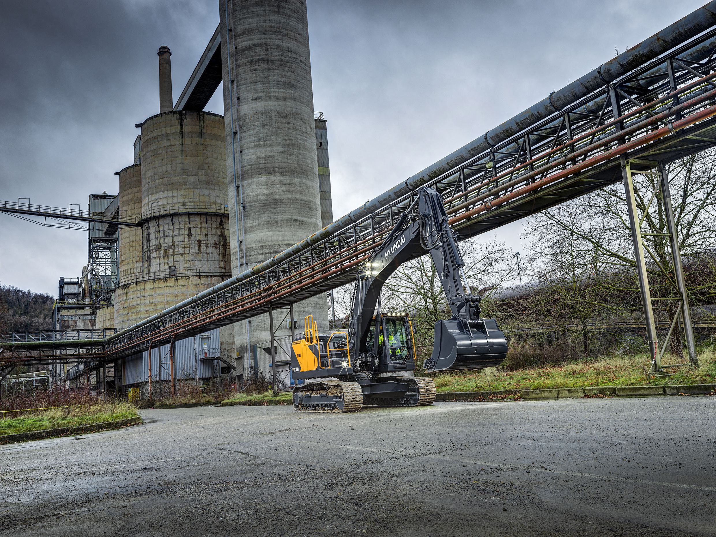 A Hyundai HX300L crawler excavator stationary on a hard-standing area at an industrial site, with large concrete silos and a conveyor system visible in the background.