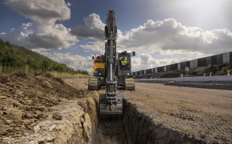 A Volvo CE tracked excavator digging a narrow trench on an open construction site, with an operator visible in the cab, a road barrier and embankment in the background, and a cloudy sky overhead