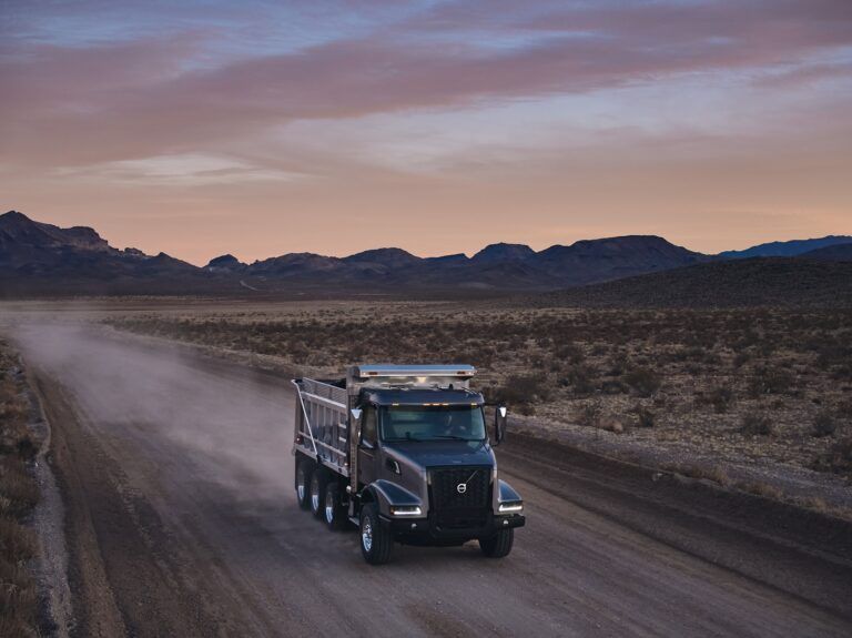 A dark grey Volvo VHD 300 dump truck driving along a dusty desert road at dusk, with mountains and a wide open landscape visible in the background.