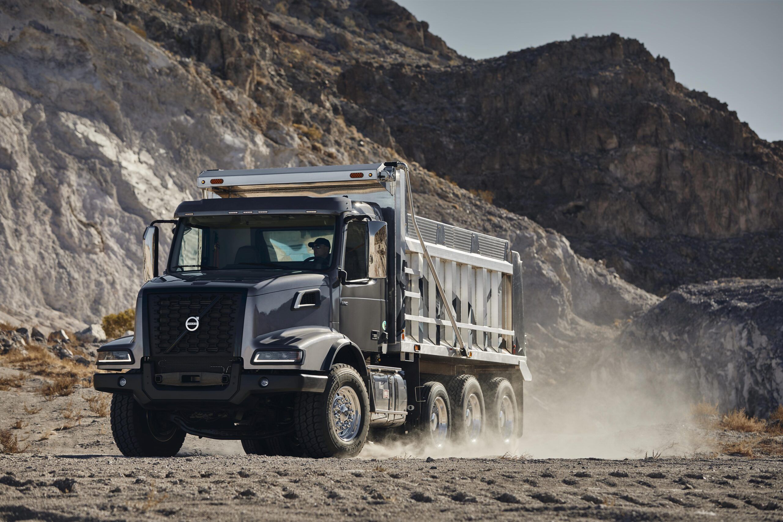 A dark grey Volvo VHD 300 dump truck with a loaded tipper body moving through a dusty quarry environment, with steep rocky terrain visible in the background and a driver visible through the cab window.