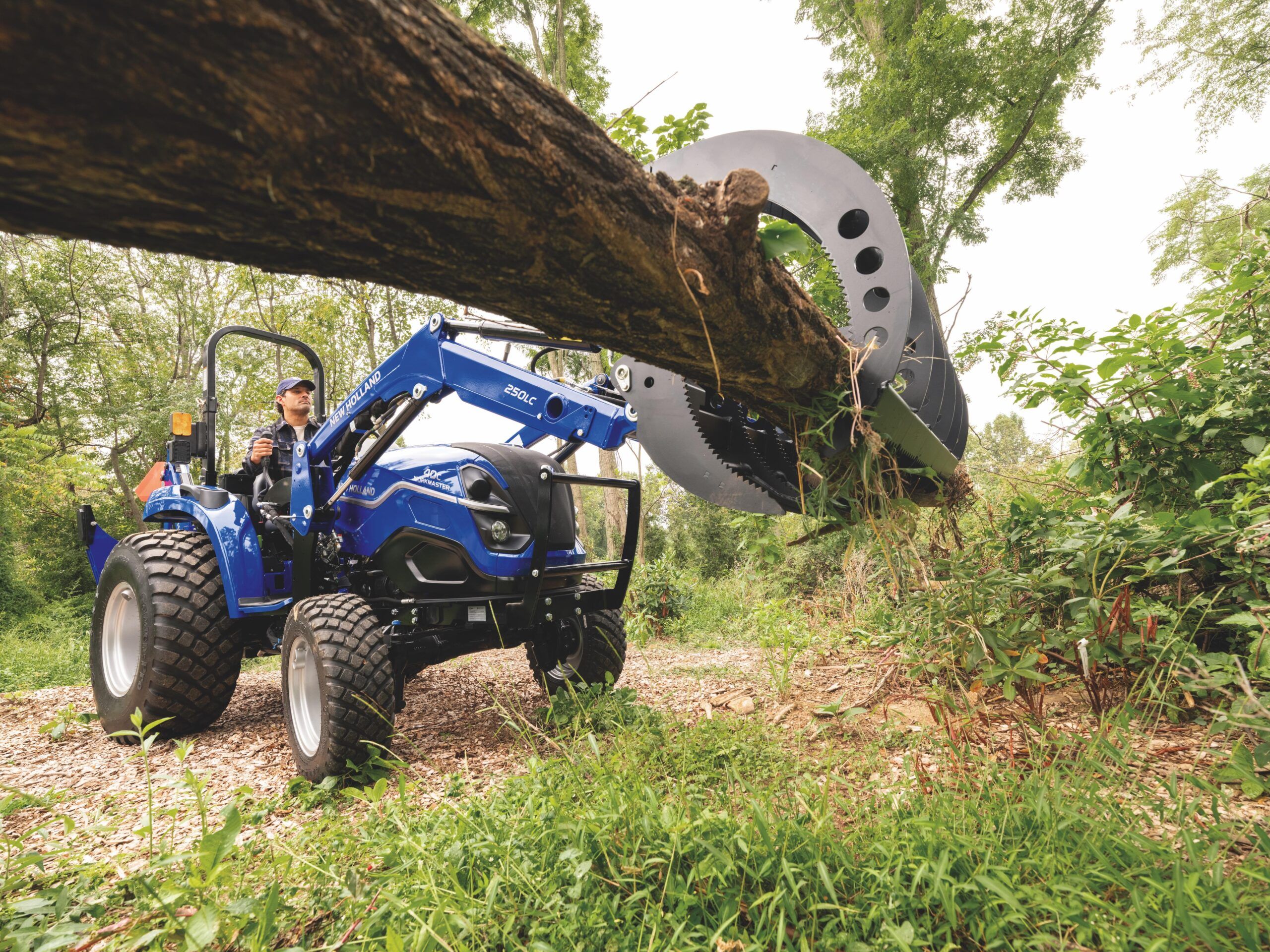 A New Holland Workmaster compact tractor with 250LC front loader carries hay in its bucket on a grassed area, with a wooden barn visible in the background. The tractor is fitted with R3+ hybrid tyres