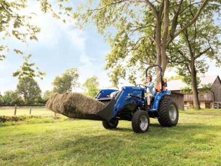 A New Holland WORKMASTER compact tractor with 250LC front loader carries hay in its bucket on a grassed area, with a wooden barn visible in the background. The tractor is fitted with R3+ hybrid tyres