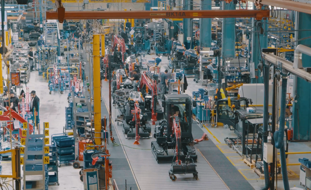 Interior view of Yanmar CE's Saint-Dizier production line, showing compact excavators in various stages of assembly with overhead cranes and workers on the factory floor.