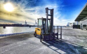 A Jungheinrich EFG 5 electric counterbalance forklift in yellow and grey, fitted with a multi-tine attachment, parked on a waterfront quayside. Port cranes and a river are visible in the background under a bright winter sky.