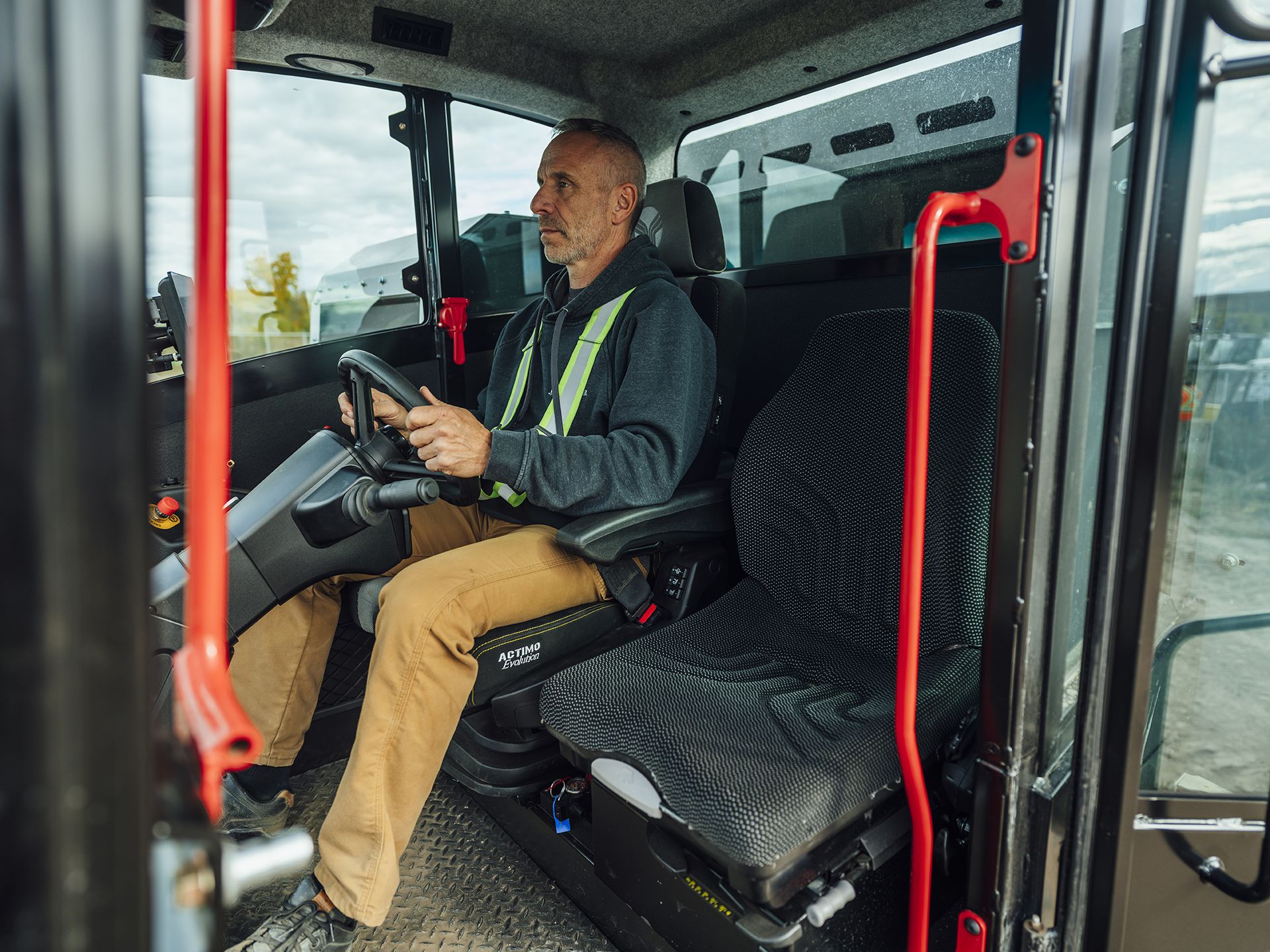 An operator seated at the controls inside the Prinoth Panther T23r cab, with a steering wheel, joystick and air-ride seat visible, viewed through the open cab door.