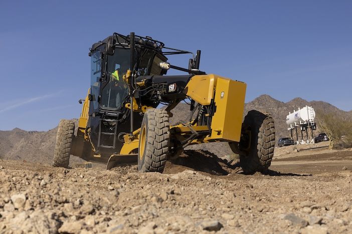 A Cat 150 motor grader working on a rocky gravel surface with desert mountains in the background. The 150 badge is visible on the side panel.