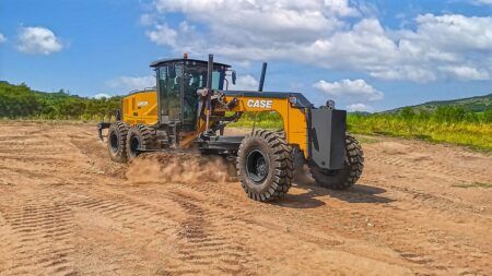 A yellow Case GR935 motor grader working on a dirt surface, kicking up dust, with green vegetation and a blue sky in the background.