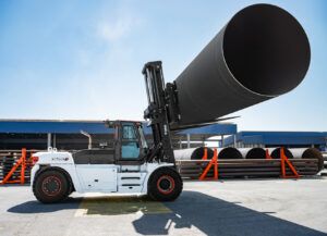 A white Bobcat DV250S-9 heavy-duty diesel forklift with red wheel rims lifts a large black steel pipe at an outdoor industrial site. Steel pipes and blue warehouse buildings are visible in the background.