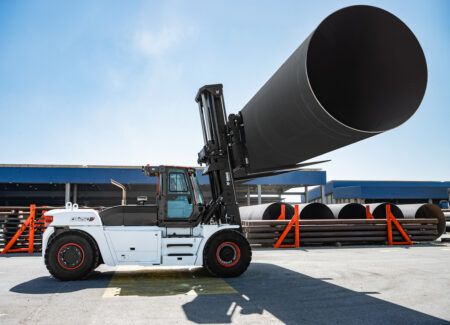 A white Bobcat DV250S-9 heavy-duty diesel forklift with red wheel rims lifts a large black steel pipe at an outdoor industrial site. Steel pipes and blue warehouse buildings are visible in the background.