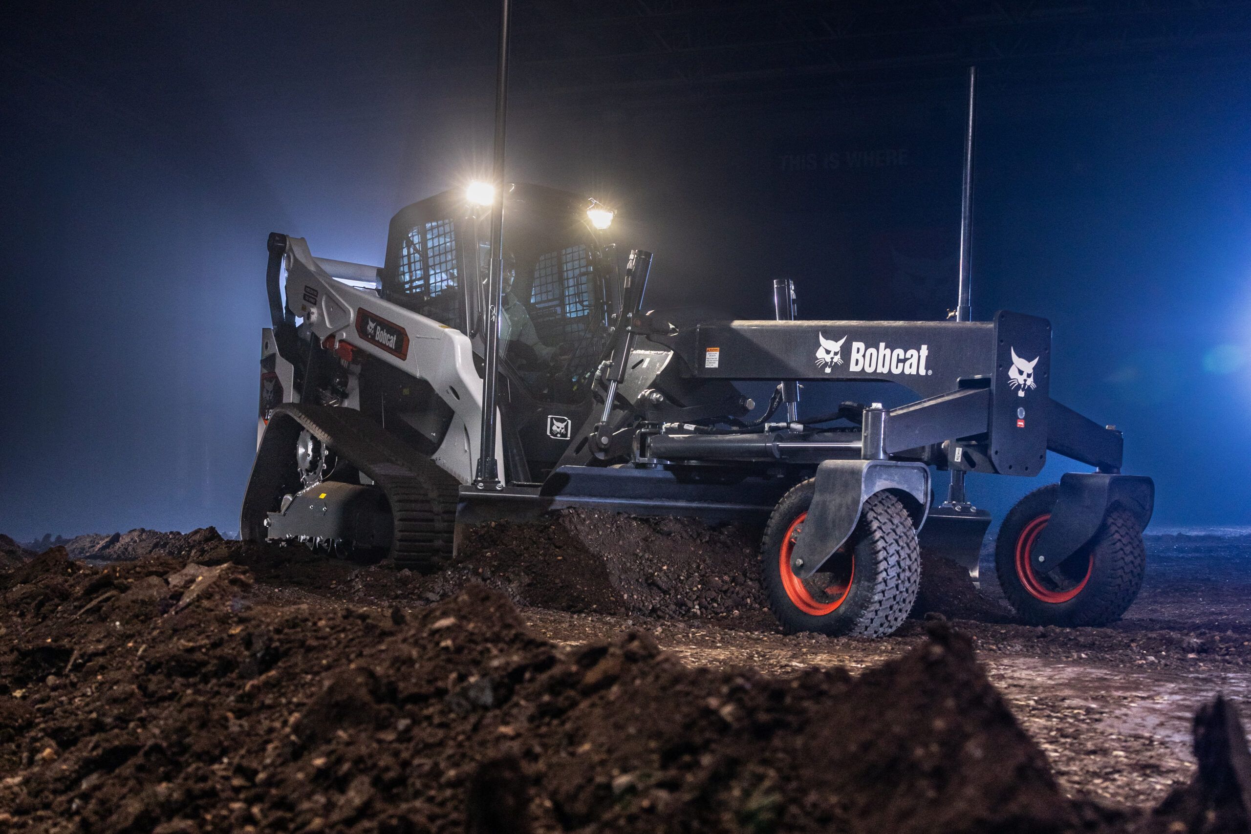 A white and black Bobcat compact track loader with cab lights illuminated, working on a dirt surface at night with a rear grading attachment fitted.