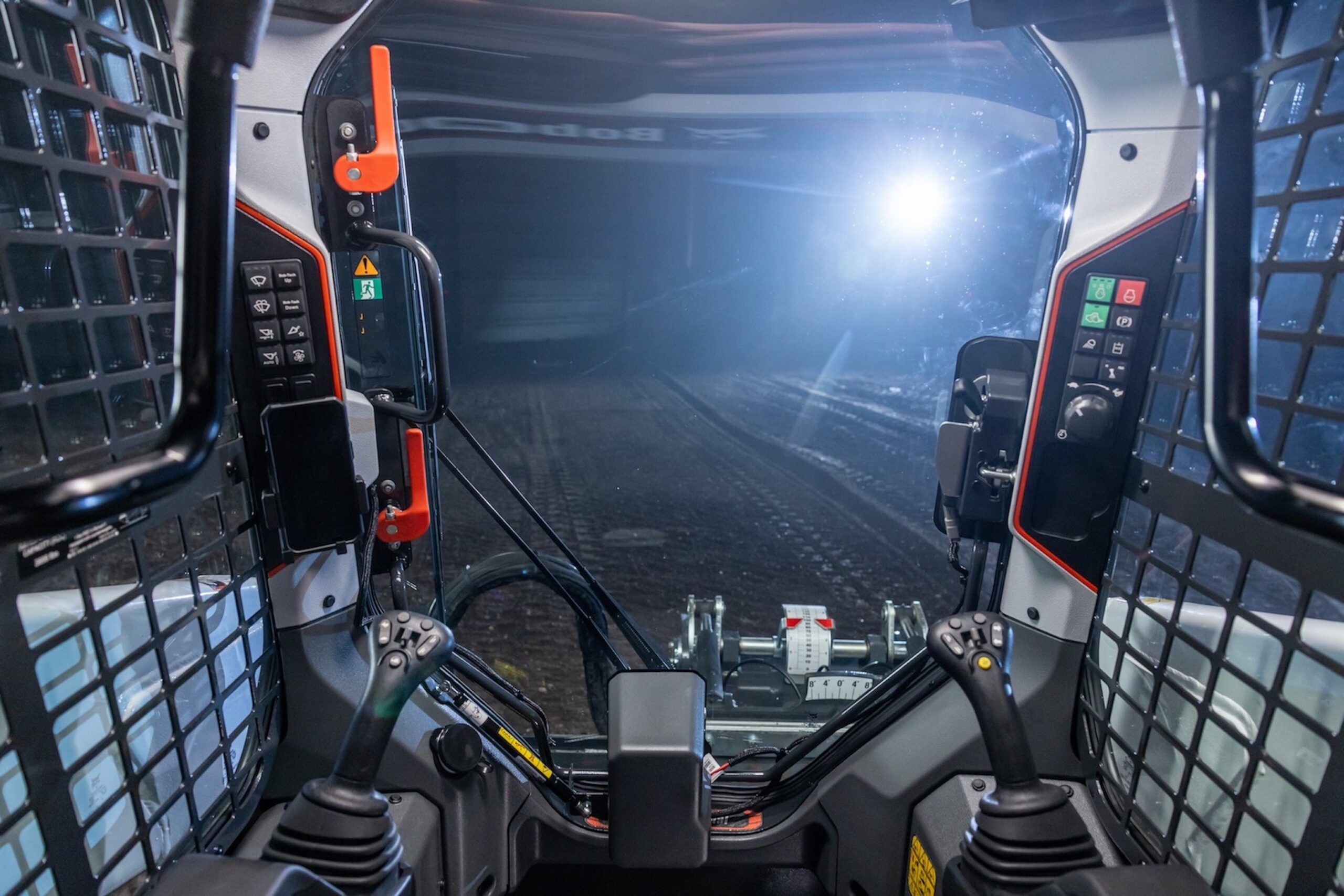 Interior view of a Bobcat R-Series compact loader cab, showing dual joystick controls, orange accent detailing, mesh side guards and a forward view through the cab door