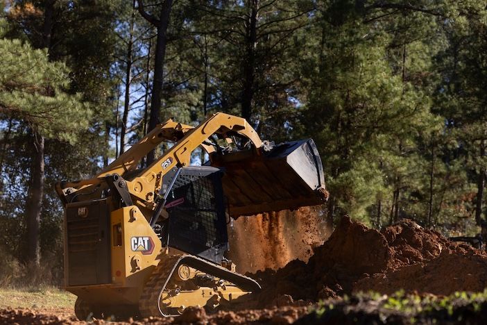 A Cat 253 compact track loader working on a dirt-moving application in a wooded outdoor setting, with the bucket raised and loaded with soil.