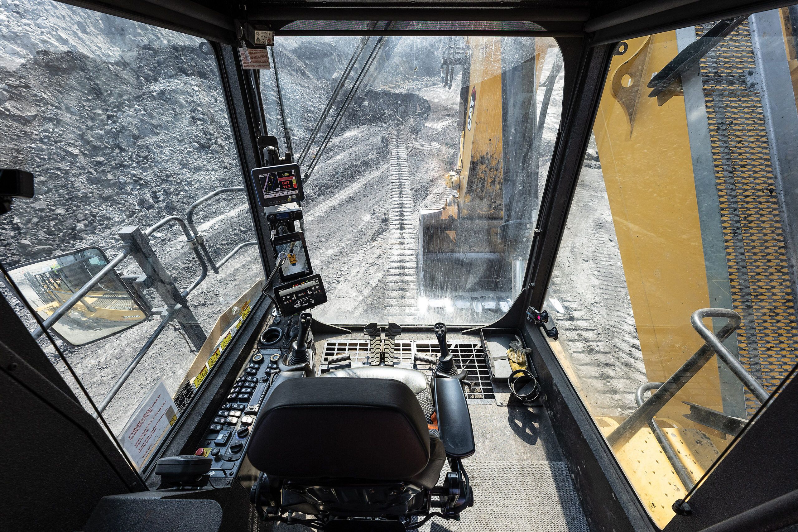 Interior view of the Caterpillar 6040 operator cab showing the ergonomic seat, dual joystick controls, dashboard-mounted touchscreen displays and large front and side windows looking out onto a mining face.