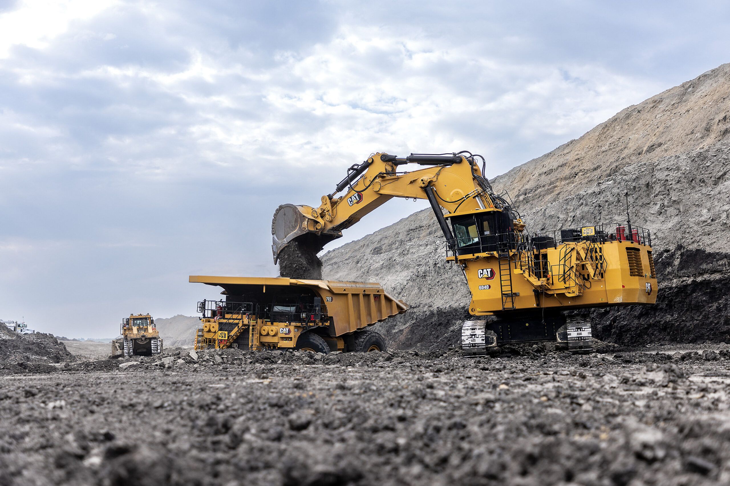 A yellow Caterpillar 6040 hydraulic mining shovel in face shovel configuration depositing material into the tray of a large Cat haul truck at a mine site, with another machine visible in the background.