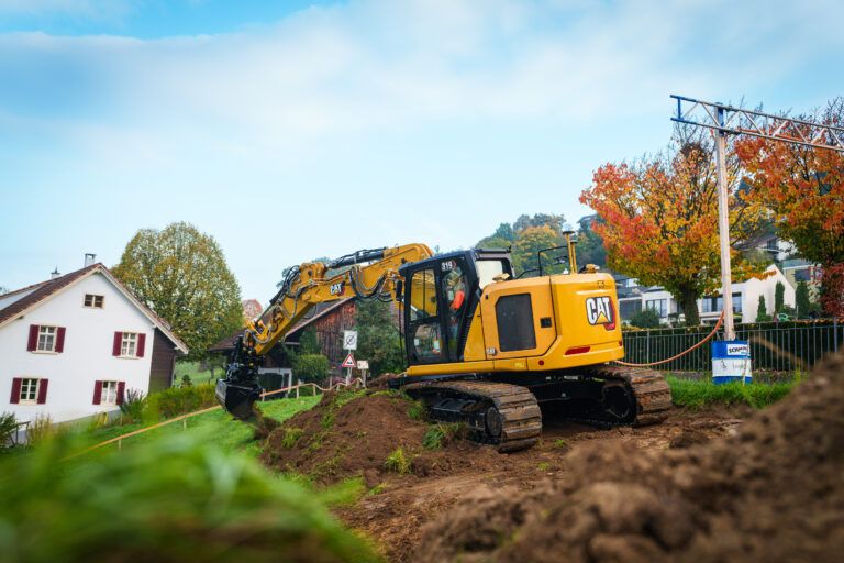 : A yellow Cat 319 crawler excavator operating on a residential construction site, digging into a mound of soil in the foreground. A white house with red shutters and autumn trees are visible in the background under a partly cloudy sky.