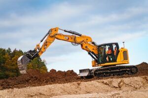 A Cat 319 compact radius excavator lifting a bucket of excavated soil on a construction site, with an operator in high-visibility clothing visible in the cab.