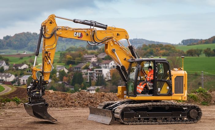 A Cat 319 compact radius excavator with tilt rotator and blade attachment working on a hillside site, with a European village and rolling green landscape in the background.