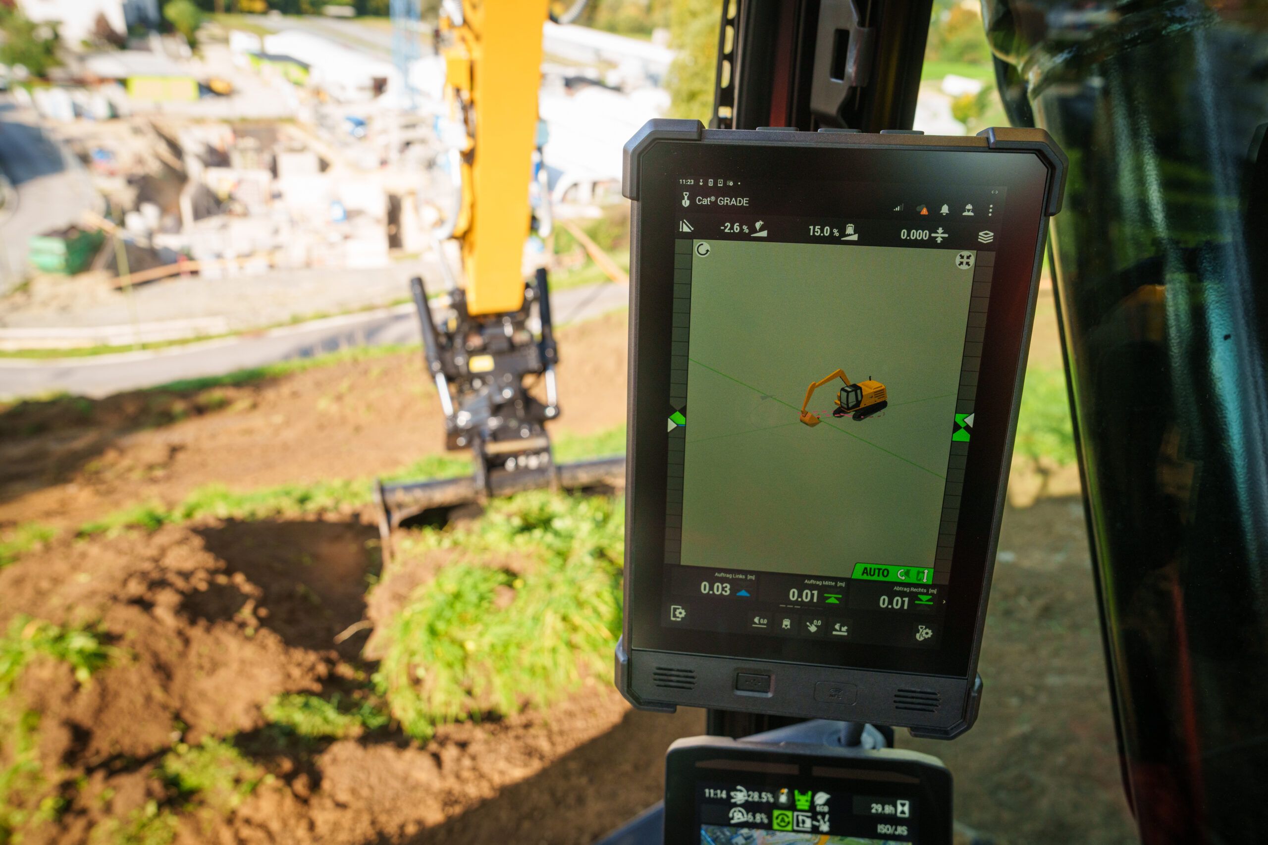 A close-up of a tablet-style Cat Grade display mounted inside an excavator cab, showing a 3D grading interface with slope and depth readings. Through the cab window in the background, the excavator's tiltrotator attachment and a construction site are visible.