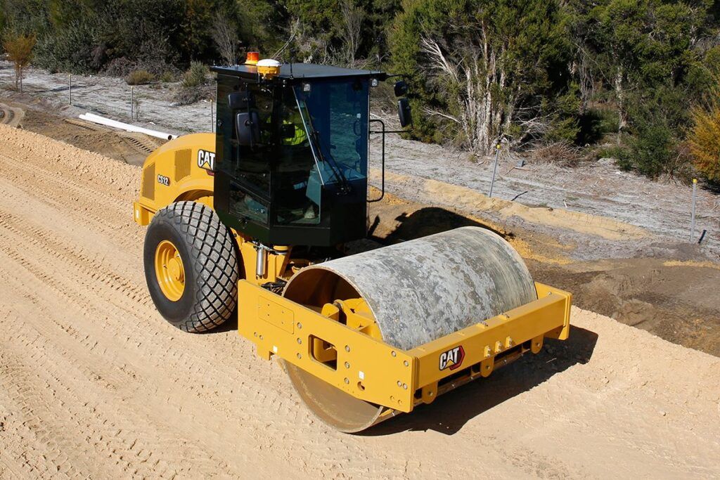 A Cat CS12 single-drum soil compactor compacts a sandy embankment on a road construction site, viewed from above 