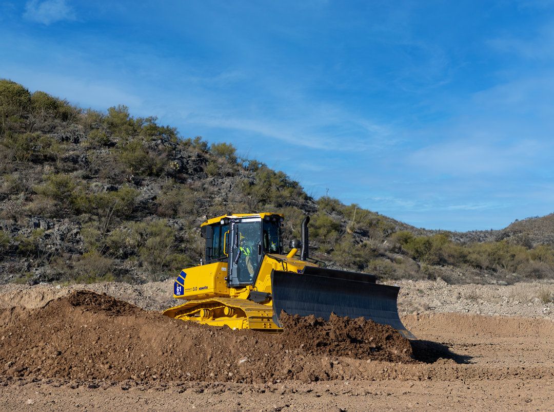 A yellow Komatsu large crawler dozer working on a dirt surface with rocky terrain and a blue sky in the background